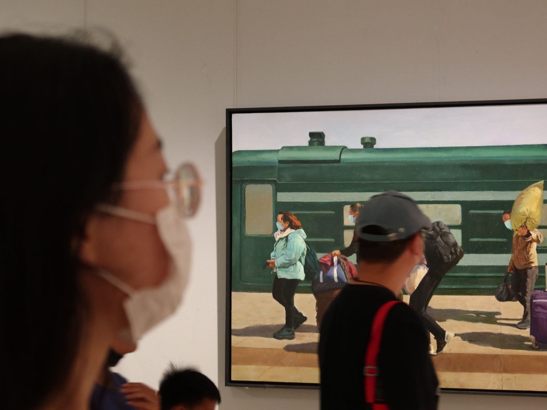 Person wearing a mask, looking at someone else who is looking at a painting of people getting off a train, all wearing masks