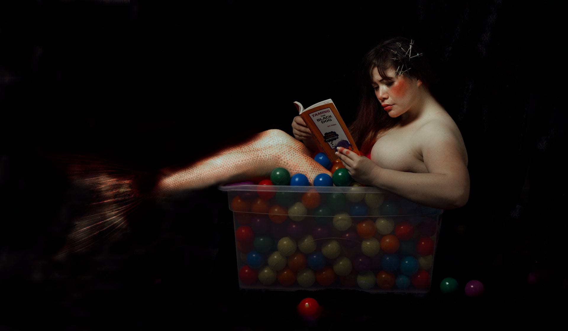 Person sitting in a bucket full of colourful balls, reading a book